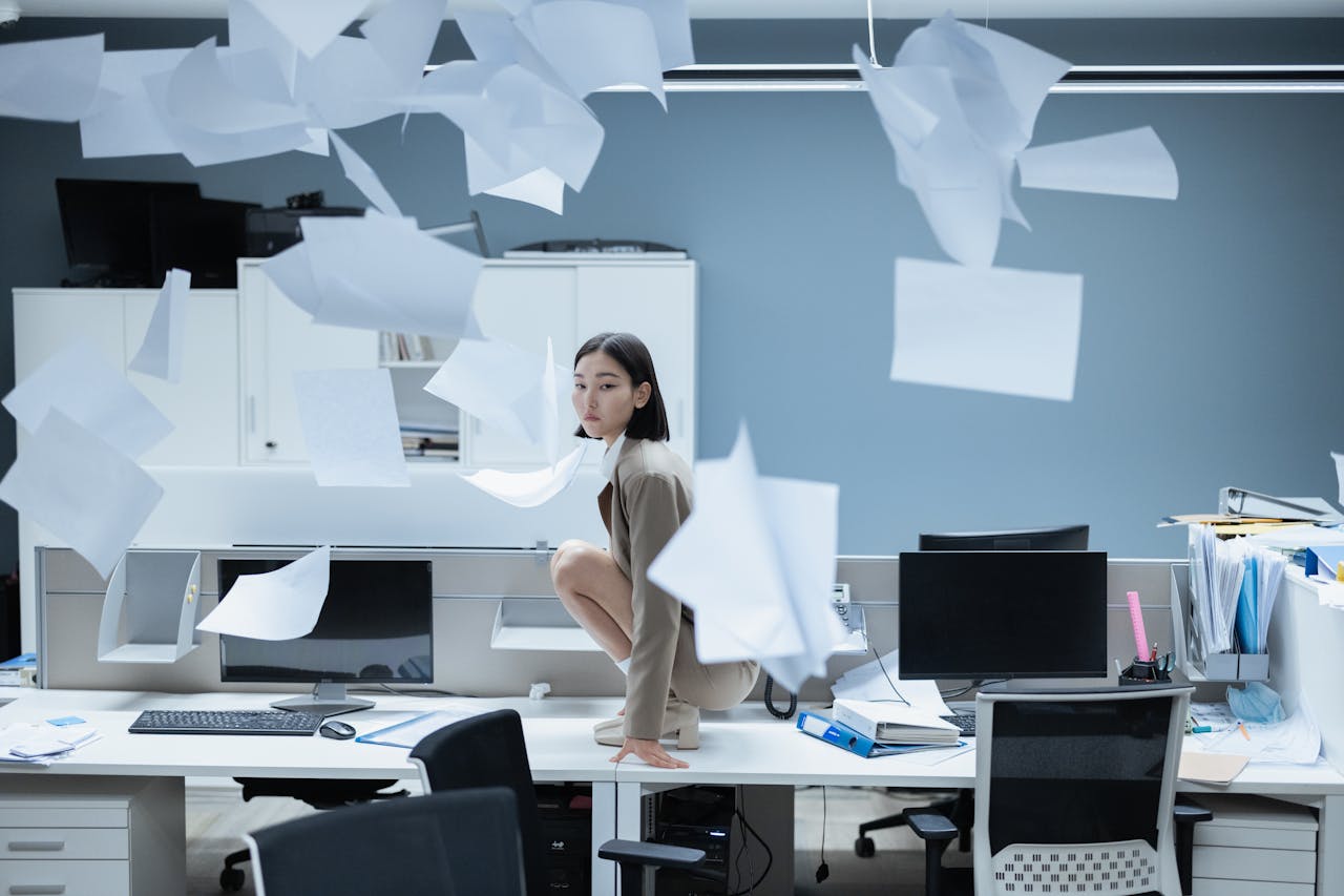 Woman perched on a desk in a chaotic office with papers flying, representing workplace stress.